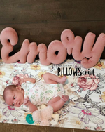 Load image into Gallery viewer, Baby lying on a floral blanket with a pink personalized PillowScript pillow spelling out 'Emory'.
