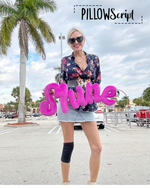 Load image into Gallery viewer, Woman holding PillowScript pillow in pink letters spelling 'Shine' in a parking lot with palm trees and a clear sky.
